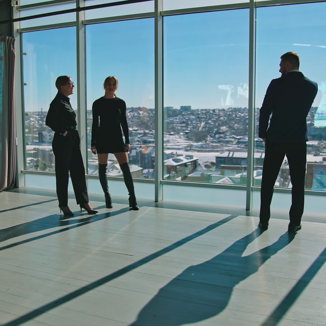 Male entrepreneur in suit with a phone and women indoors. Beautiful young female partners waving hello to a businessman standing near the window