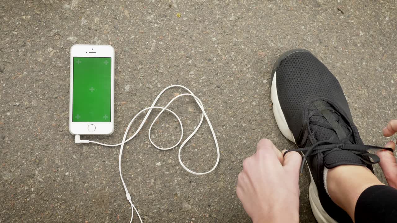 una chica deportista se ata los cordones de los zapatos mientras su teléfono inteligente y auriculares están en el suelo, pantalla de croma, concepto de comunicación