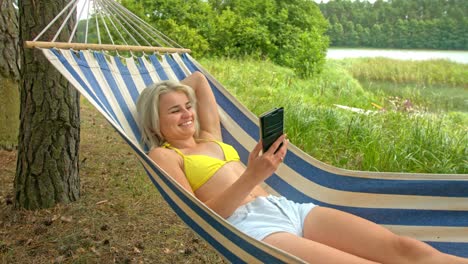 sexy young girl smiling towards her smartphone having a video chat while lying on a hammock