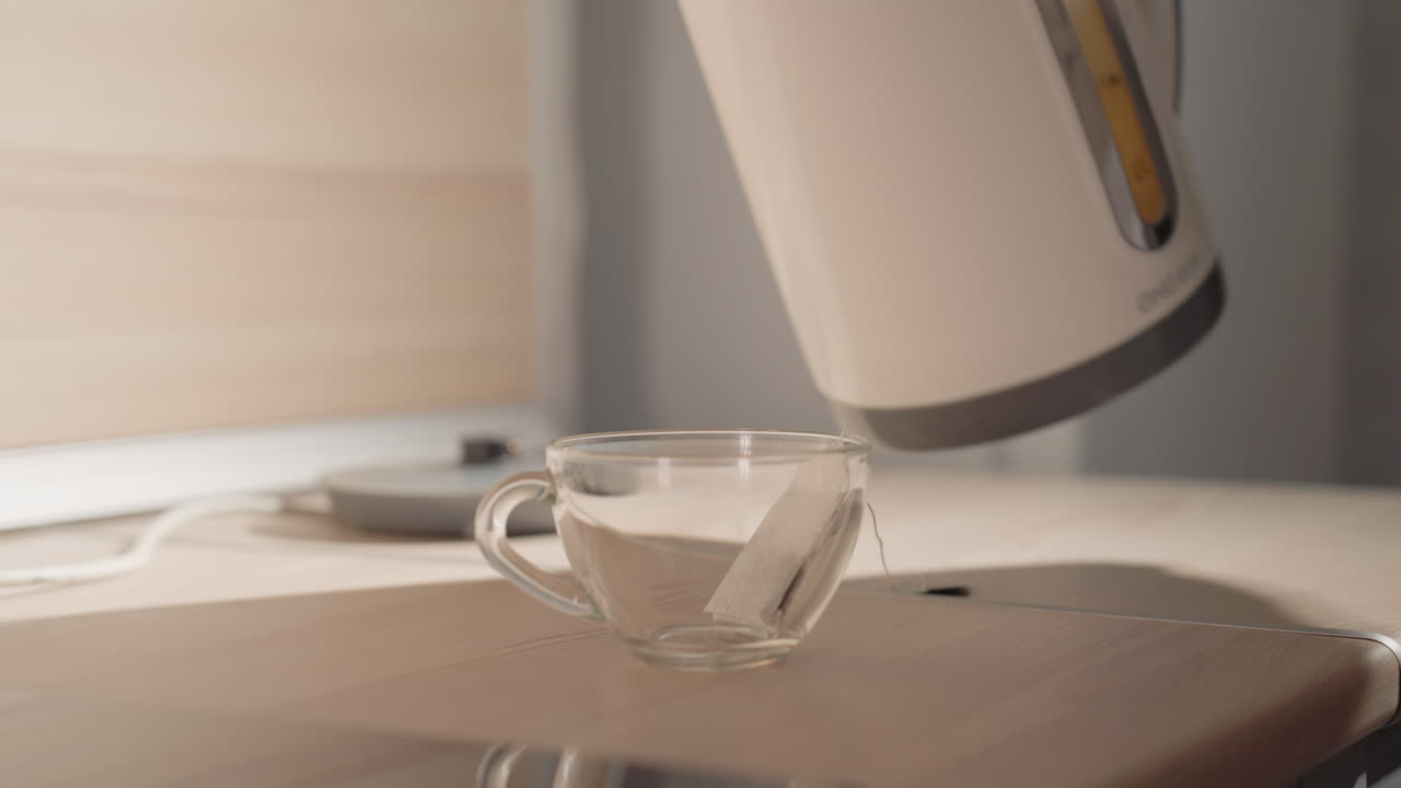 Close-up of hot water being poured from electric kettle into clear glass cup with tea bag. Soft light creates warm, serene atmosphere in modern minimalist kitchen