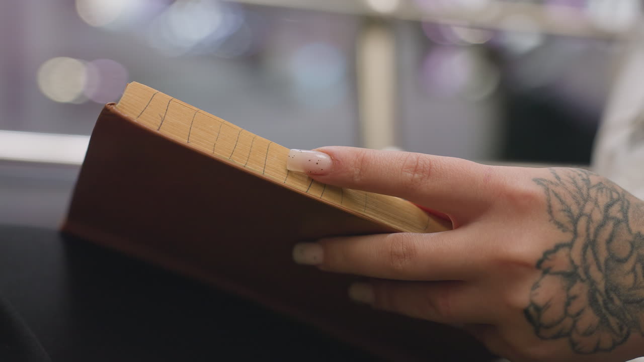 Close up of lady hand with fixed nails and floral tattoo gently flipping through book pages in soft indoor light, creating peaceful reading atmosphere with blurred background