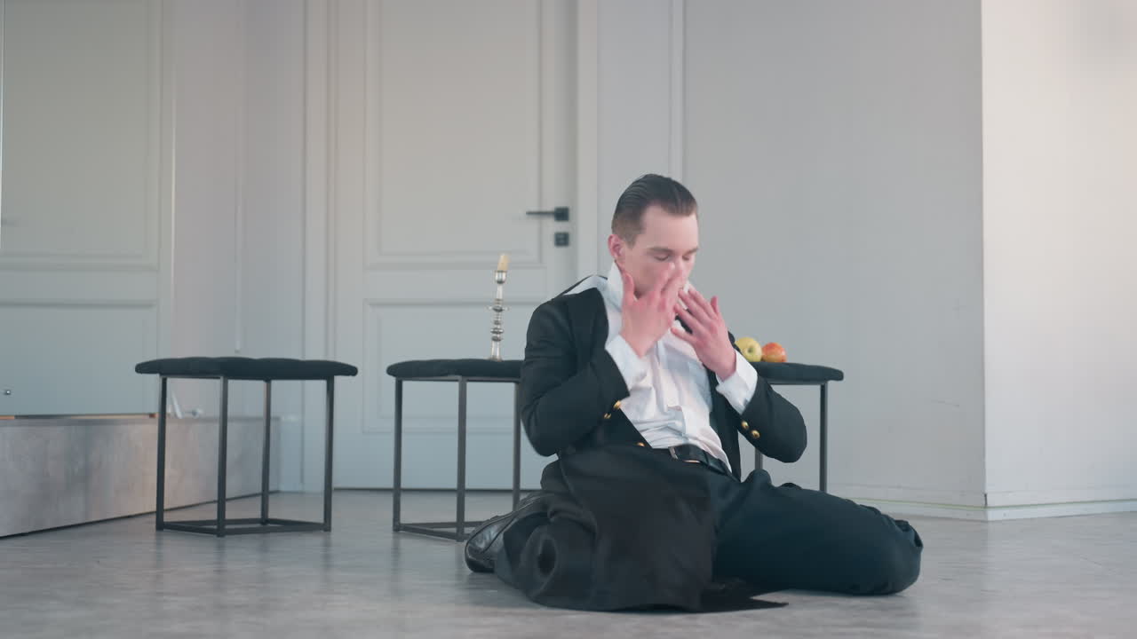 dramatic dancer in black coat and white shirt performs expressive ground movement beside wall mirror with apples on stool and candle stand, illuminated by soft studio light