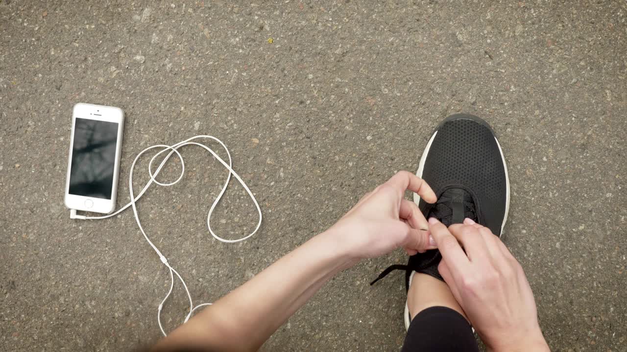 Ragazza sportiva si allaccia le scarpe mentre il suo smartphone e gli auricolari sono a terra, concetto di comunicazione