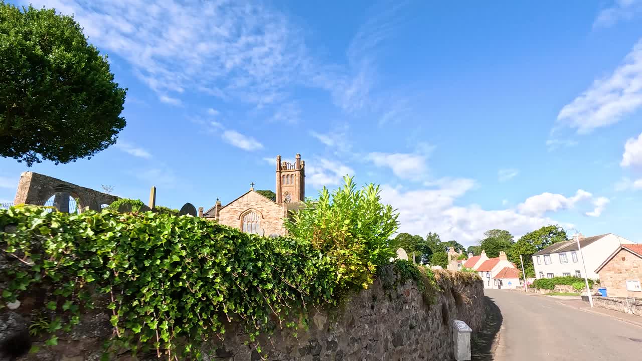 Daytime street-level walk past stone church, cemetery, stone wall, village houses, and blue sky