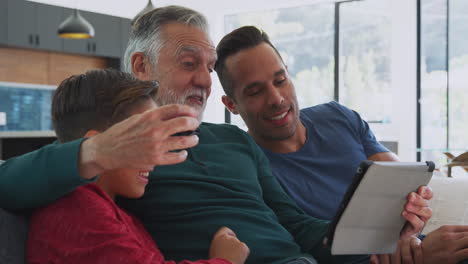 multi-generation male hispanic family sitting on sofa at home watching movie on digital tablet