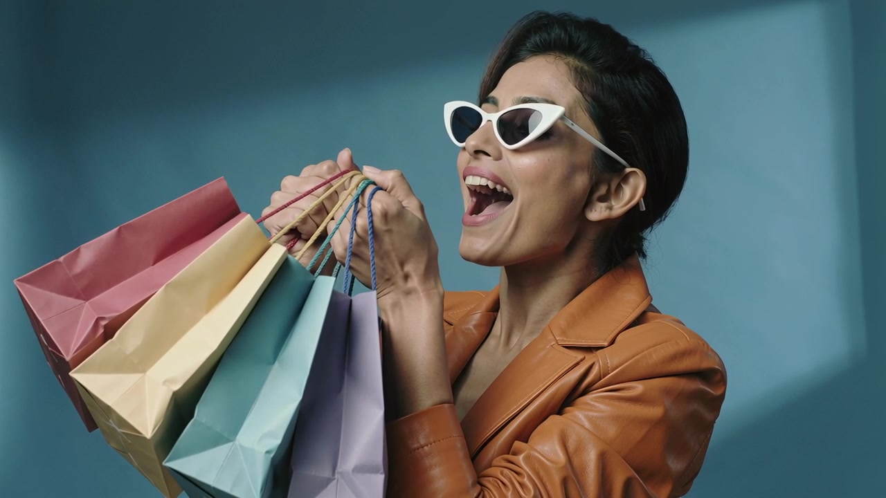 Una donna che solleva le borse portando i manici alla bocca ridendo, con occhiali da sole cat-eye e blazer marrone