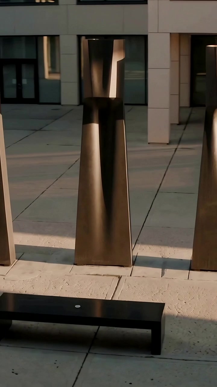 Video verticale: La telecamera inizia a panoramare attraverso una piazza vicino a un edificio, mostrando colonne di bronzo e una panchina.