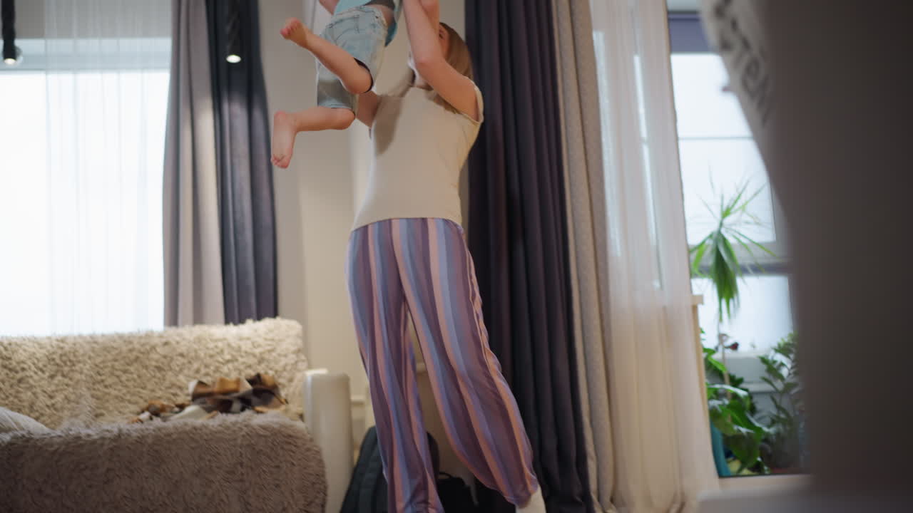 Eine Mutter hebt ihren kleinen Sohn in einem spielerischen Moment der Verbundenheit in einem hellen, gemütlichen Wohnzimmer hoch in die Luft und drückt Freude, Spaß, Zuneigung, Energie und Wärme in einer fröhlichen, unbeschwerten Familieninteraktion zu Hause aus.
