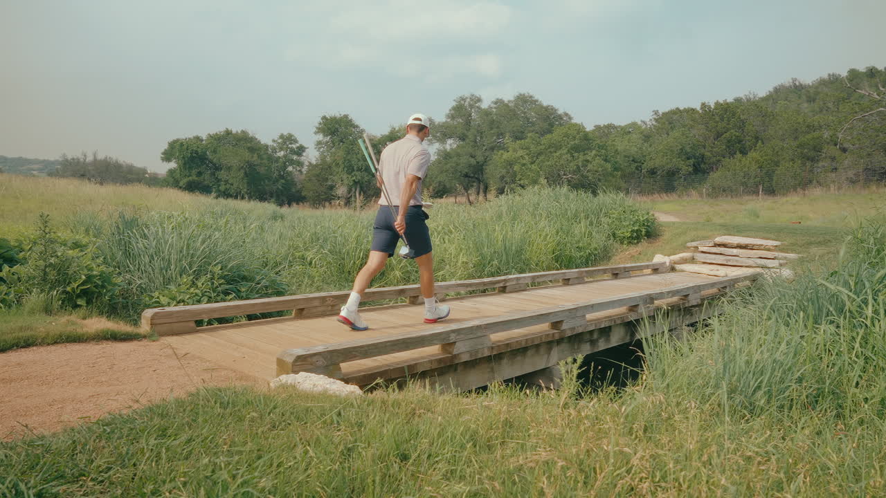 A male golfer walks across a scenic bridge carrying multiple clubs to the tee box in slow motion, captured from the side. The shot highlights his calm, focused preparation on the course.
