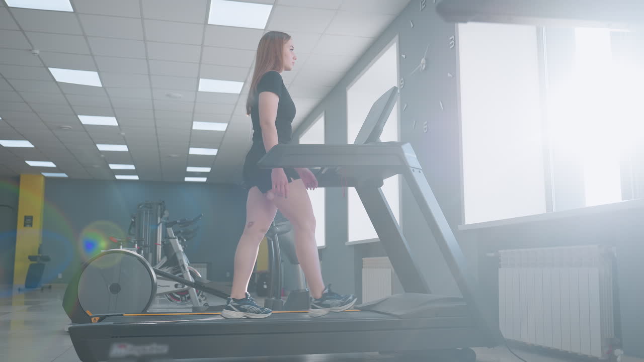 athlete walking on treadmill with focus as sunlight filters through large window creating lens flare in modern gym with exercise bikes elliptical trainer tiled floor and gray wall clock visible