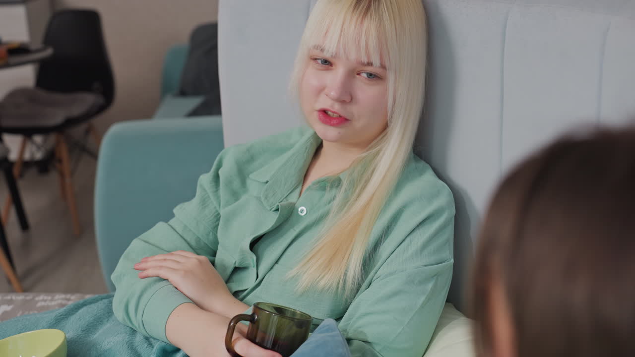 Blonde girl in green shirt reclines against headboard, holding cup and chatting with unseen friend. Indoor setting with soft furnishings suggests casual, comfortable conversation