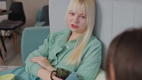 blonde girl in green shirt reclines against headboard, holding cup and chatting with unseen friend. indoor setting with soft furnishings suggests casual, comfortable conversation