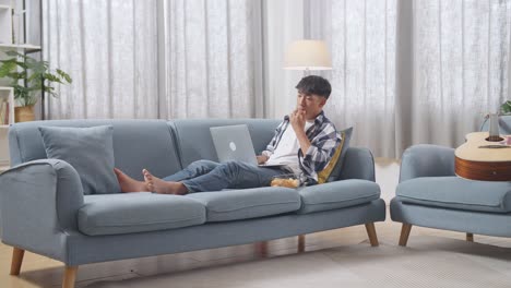 full body of asian teen boy eating snacks and typing on a laptop while lying on sofa in the living room at home