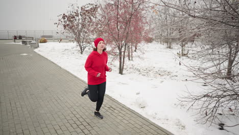 woman jogging along paved snow-covered path with a warm smile during winter, surrounded by serene park scenery, barren trees, and benches, with someone walking in the distant background