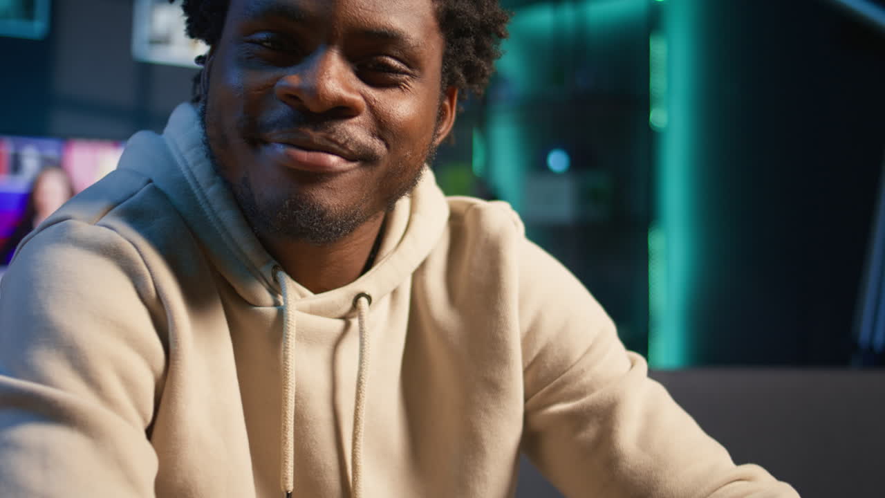 Black remote worker enjoying flexibility and comfort while browsing online on laptop in his stylish apartment decorated with neon lights. Freelance work perfectly captured in a cozy home. Camera B.