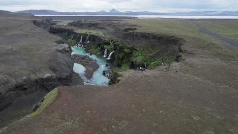 an aerial view of tourists observing a canyon with waterfalls and blue water in the icelandic highlands. the group stands along the edge, admiring the rugged landscape.