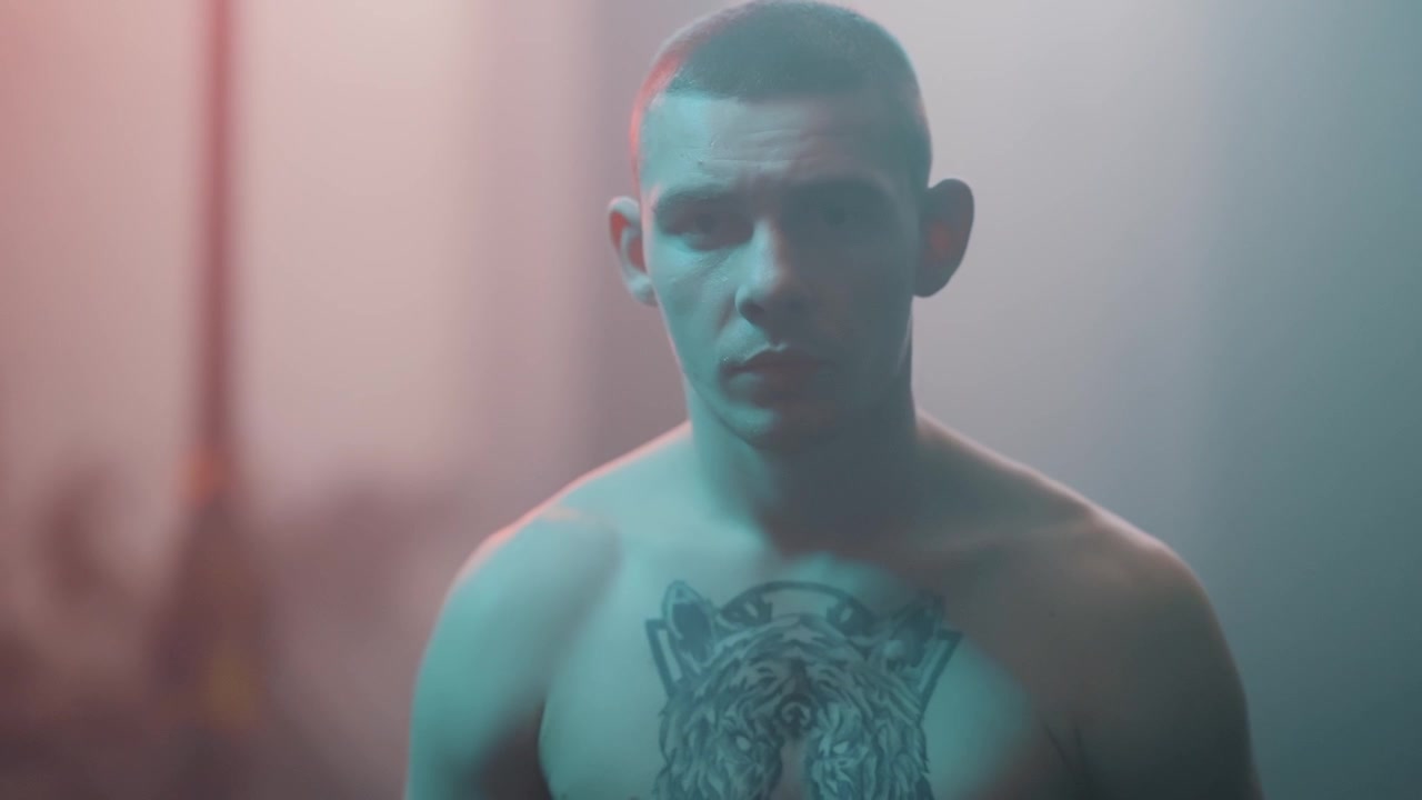 Giovane uomo serio con tatuaggio sul petto che guarda la fotocamera con illuminazione drammatica