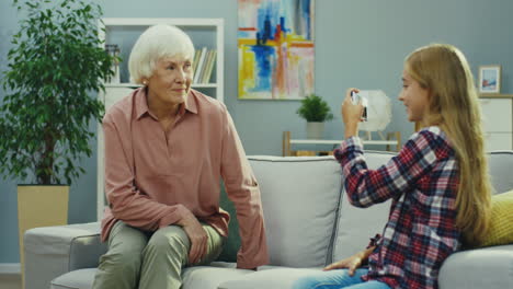 good looking old gray haired woman posing while sitting on the sofa in the room and her pretty teen granddaughter taking photos of her on the smartphone camera. portrait shot. indoor.