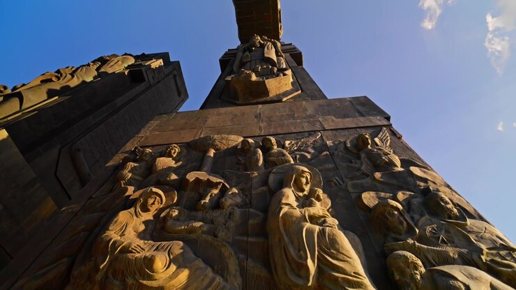 Sguardo rivolto verso un rilievo in bronzo illuminato dal sole raffigurante la Natività con la Madonna, il bambino e figure bibliche alla base di un'imponente colonna delle Cronache della Georgia