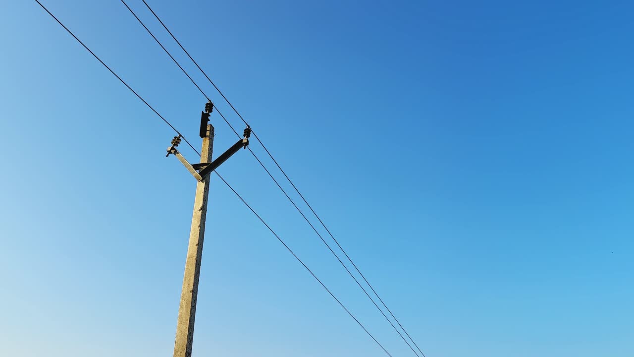 Static wide shot of a single utility pole carrying overhead power lines, isolated against a clear blue sky, emphasizing minimal composition, scale, and linear infrastructure design