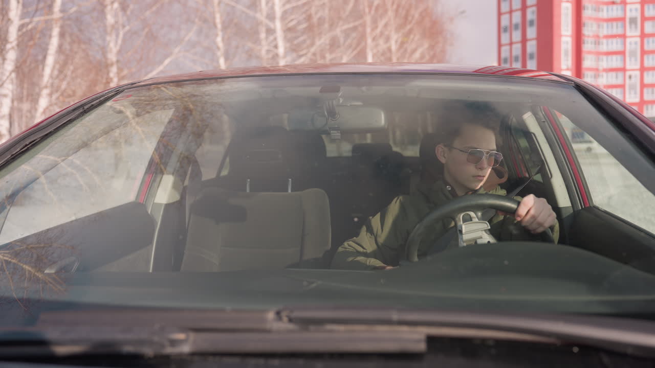 teenager wearing winter jacket seen through windshield sitting in parked car holding steering wheel with left hand while right hand rests, seatbelt unfastened, red building and snowy landscape