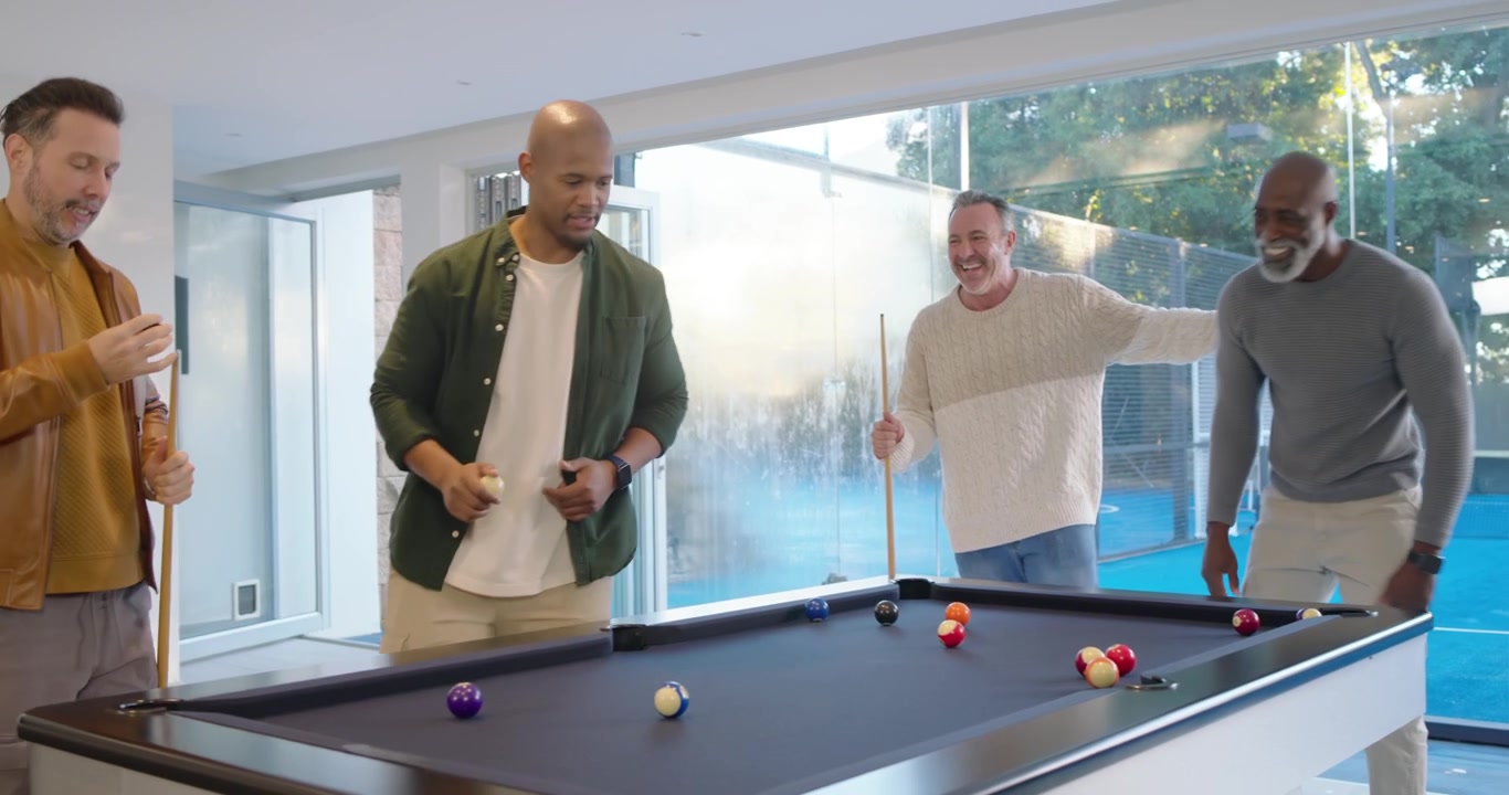 Diverse male friends laughing after initial shot, one lining up shot on black pool table