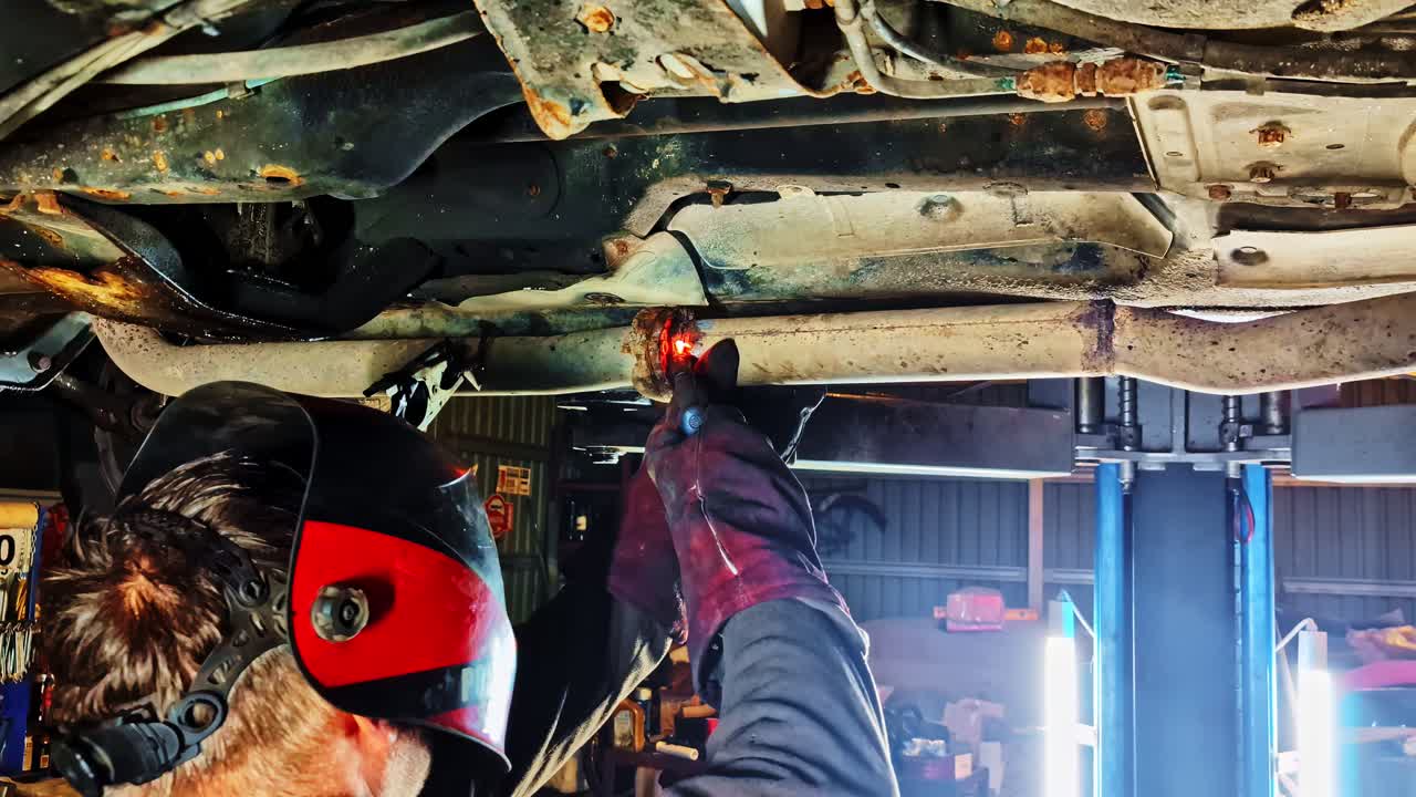 Welder sparks fly under raised car as mechanic repairs axle inside dim auto workshop