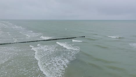 Vista-Aérea-De-Un-Antiguo-Muelle-De-Madera-En-La-Costa-Del-Mar-Báltico,-Día-De-Invierno-Nublado,-Playa-De-Arena-Blanca,-Postes-De-Madera,-Olas-Golpeando-La-Costa,-Gran-Tiro-De-Drones-Avanzando