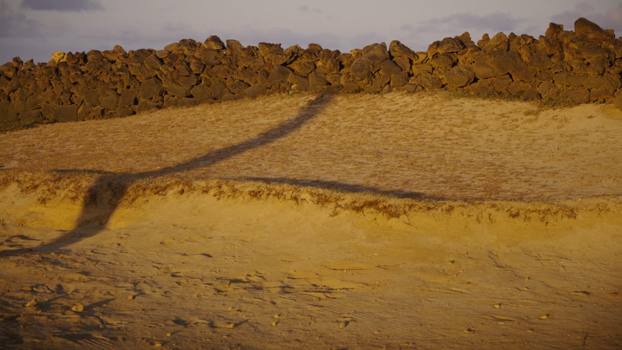 A long diagonal human shadow extends across sandy ground in the golden light of sunrise, creating a minimalist and conceptual scene with warm earthy tones