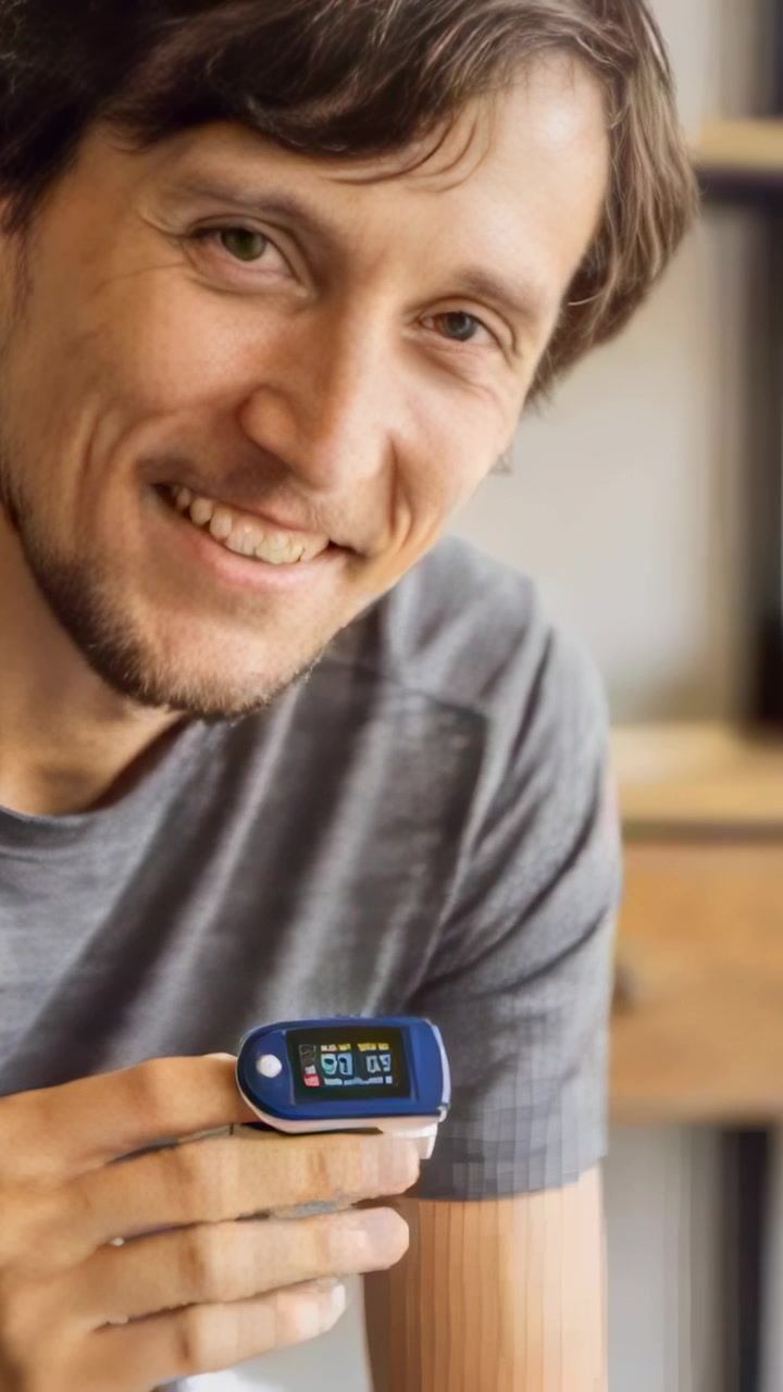 Vertical video: Smiling man in gray tee holding blue pulse ox at home showing readings to camera