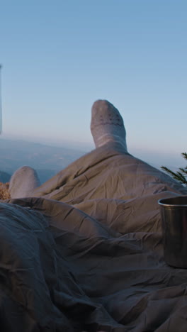 romantic hiker couple lie in tent on mountain hill and drink hot tea. two tourists rest under blanket during adventure vacation. backpacker family warm up at cold windy weather and admire the scenery.