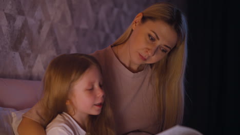 Woman-looks-at-daughter-reading-book-on-bed-at-night