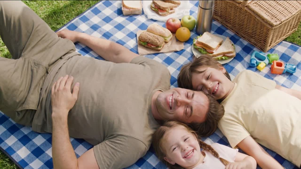 Glad familie ligger sammen på et picnictæppe med madkurv og nyder et solrigt øjeblik udendørs.