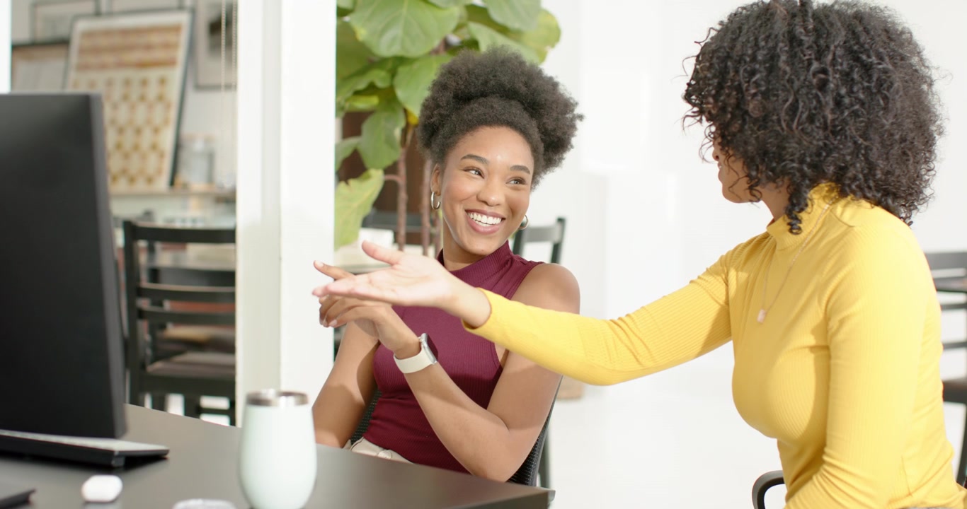 Diverse female coworkers left woman gesturing at monitor, right woman pointing and guiding at desk