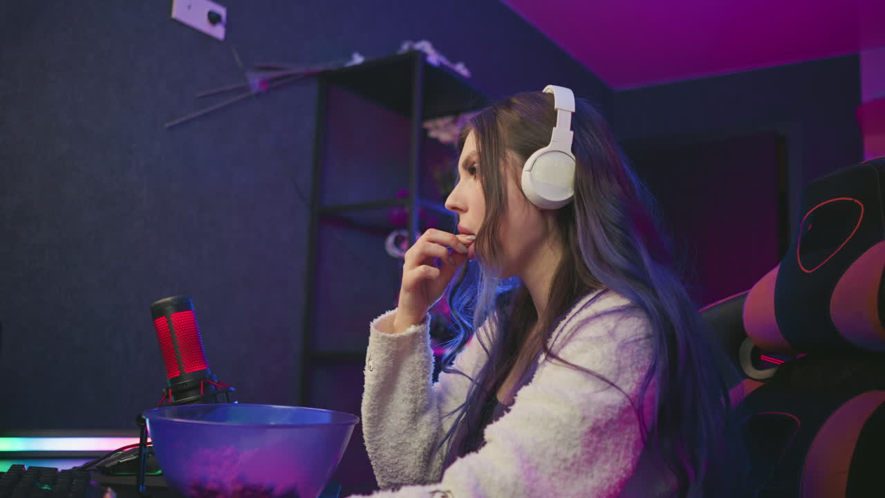 Young woman wearing headphones eats popcorn from bowl while working at computer desk in vibrant studio setup with bright LED lighting and microphone