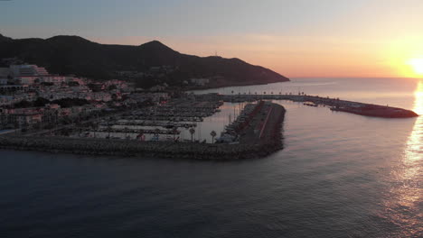 Sunrise-drone-shot-with-yacht-port-surrounded-by-calm-ocean-waves-near-coastline-town-with-mountain-range-backdrop