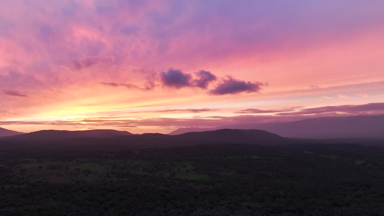 Drone lateral pan in a valley at dusk, with distant mountains and a sky full of colors—orange, yellow, blue, red, and violet. Clouds reflect rays, creating light lines as the day fades to night.