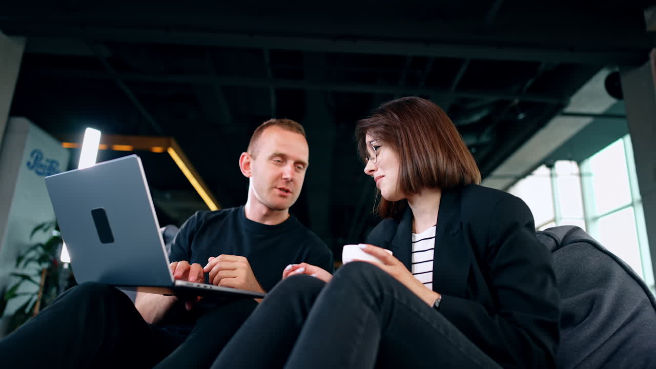 Two people sit in the bean chairs looking at laptop. Man and woman discuss lively what they see on the screen. Low angle view.