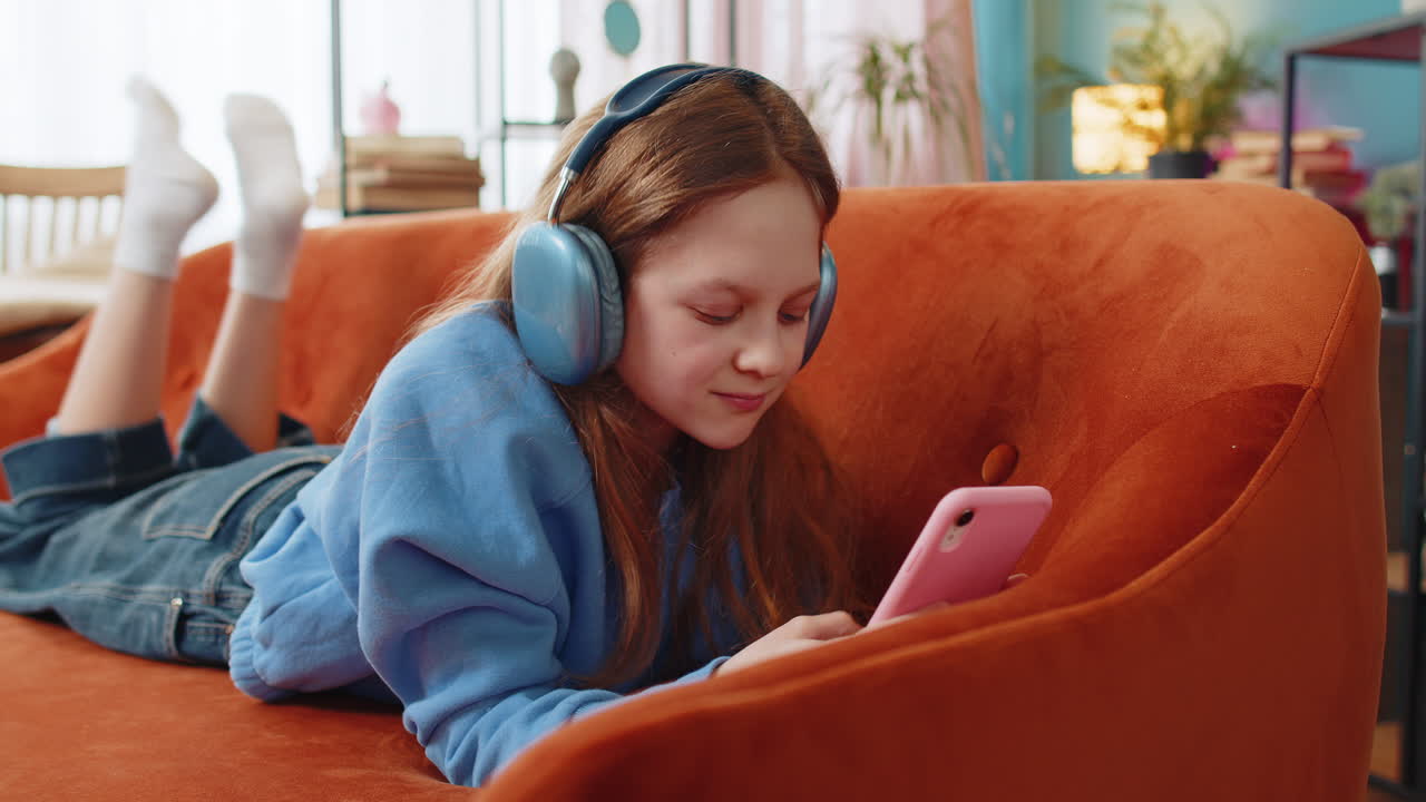 feliz niña caucásica niño en auriculares inalámbricos relajándose acostado en el sofá en casa escuchando música