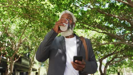Hombre-Mayor-Afroamericano-Sosteniendo-Un-Teléfono-Inteligente-Bebiendo-Café-Mientras-Caminaba-En-Un-Parque-Corporativo