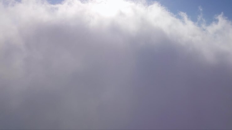 Drone ascending view through white clouds towards a bright sunlight shining