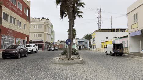 a cobblestone intersection in sal rei, cape verde, featuring a central palm tree, parked vehicles, and colorful buildings. the scene blends urban charm with a vibrant, tropical setting