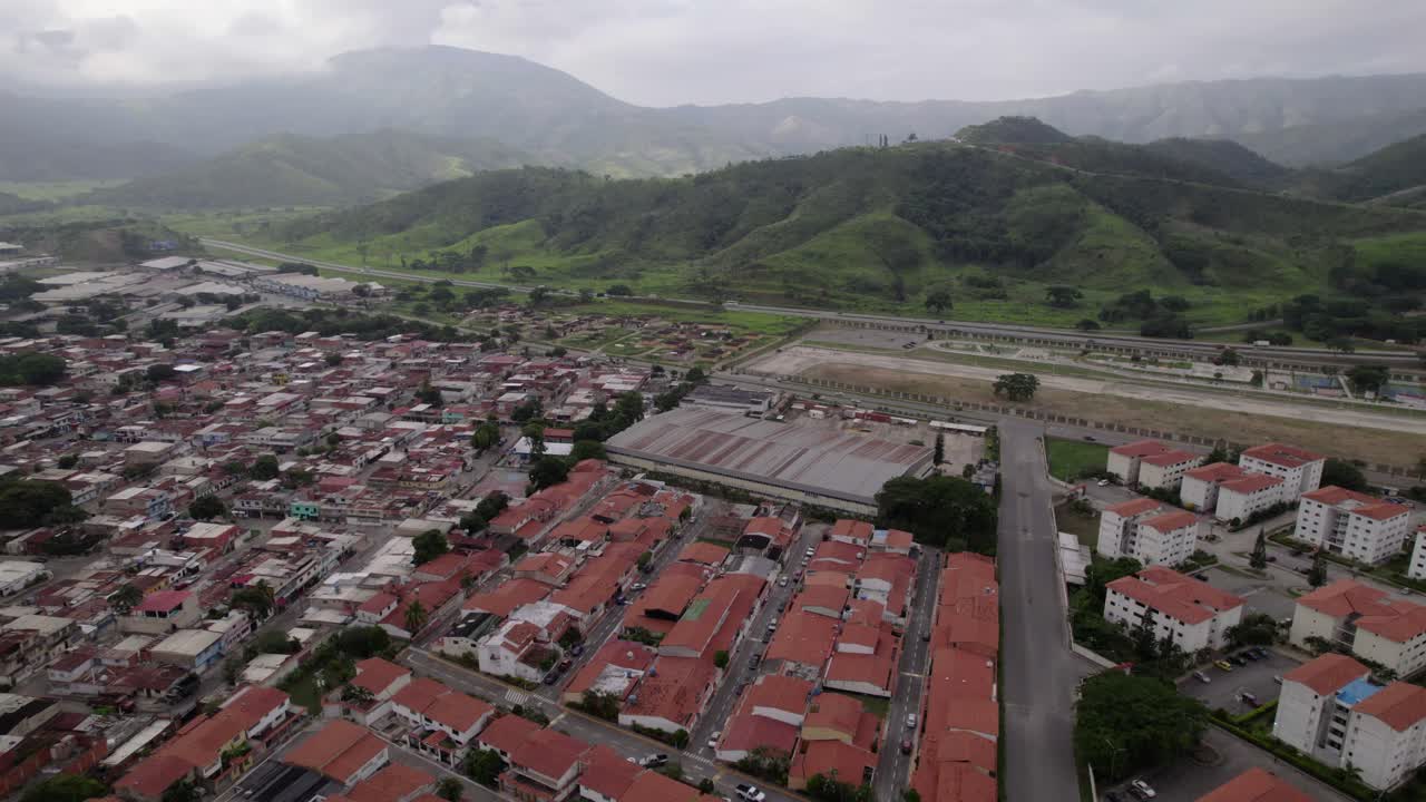 Drone shot sprawling urban and industrial development of La Victoria City, dense residential area with red-tiled roofs meet green mountains, contrast between human expansion and untouched nature