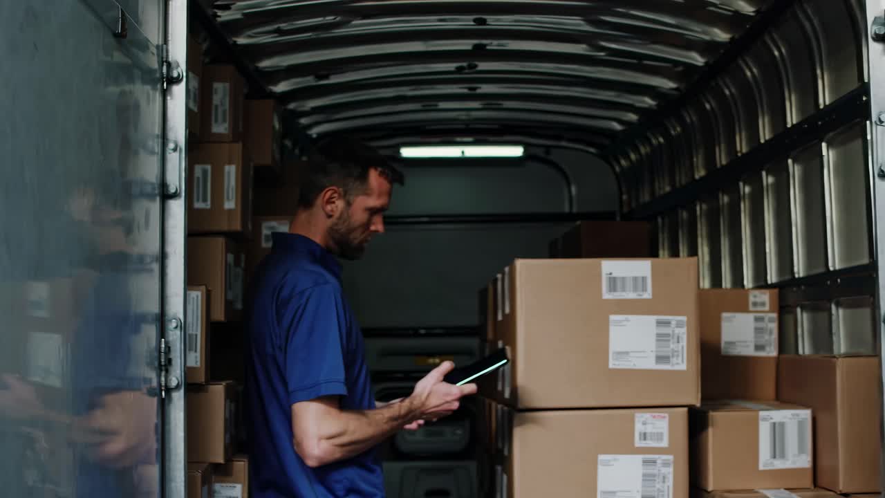 A man in a delivery truck checks packages using a tablet. Side angle captures the organized boxes