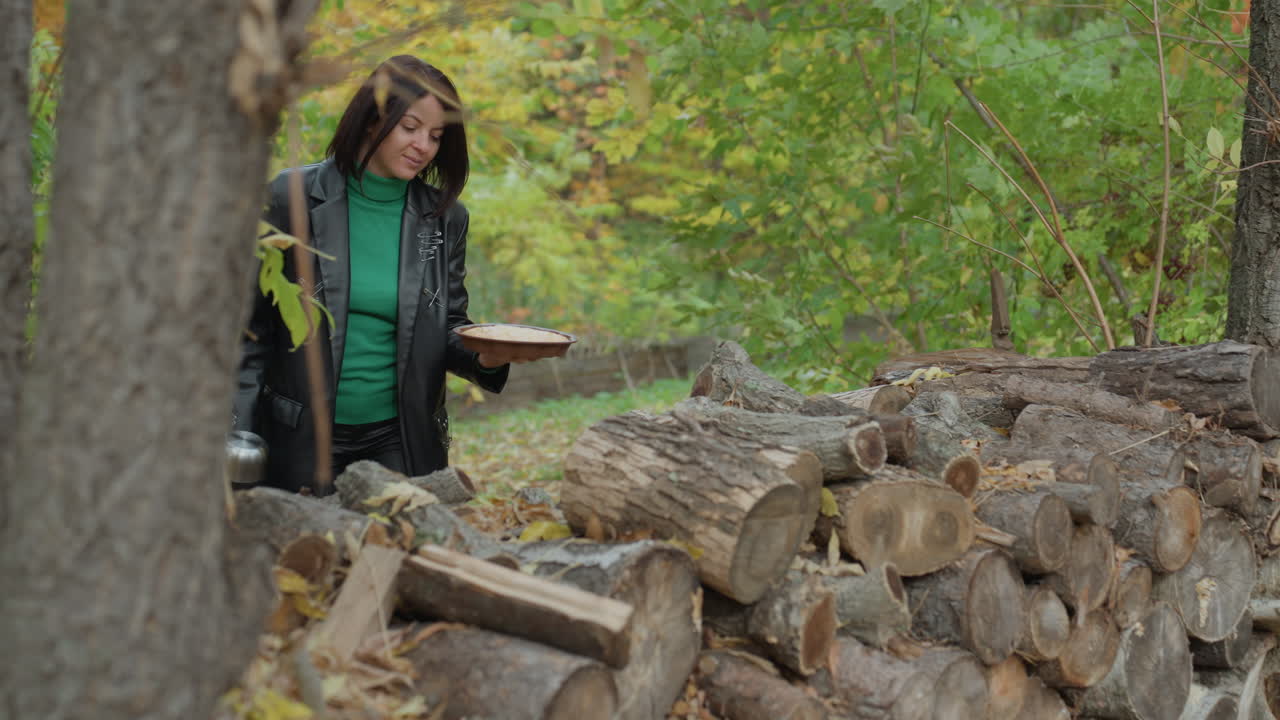 woman in leather jacket carrying thermos and small box, walking through green forest toward stacked logs, pauses at woodpile to inspect and lift log, autumn leaves and damp bark, contemplative edgy
