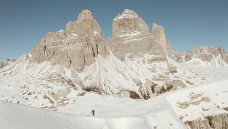 Dolly-forward-drone-shot-of-trekker-walking-along-a-steep-snowy-ridge-dolomites-tre-cime-in-the-background