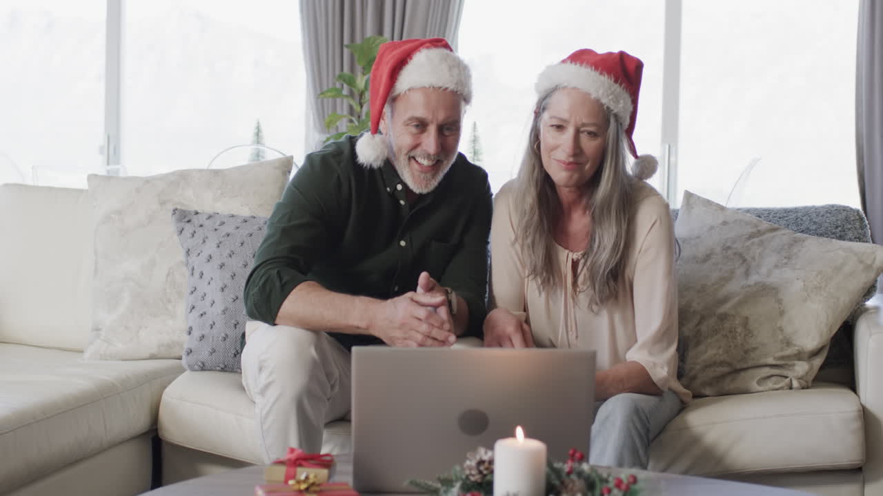 una pareja caucásica de mediana edad con sombreros de papá noel en un ordenador portátil hablando por video en navidad en casa, en cámara lenta.