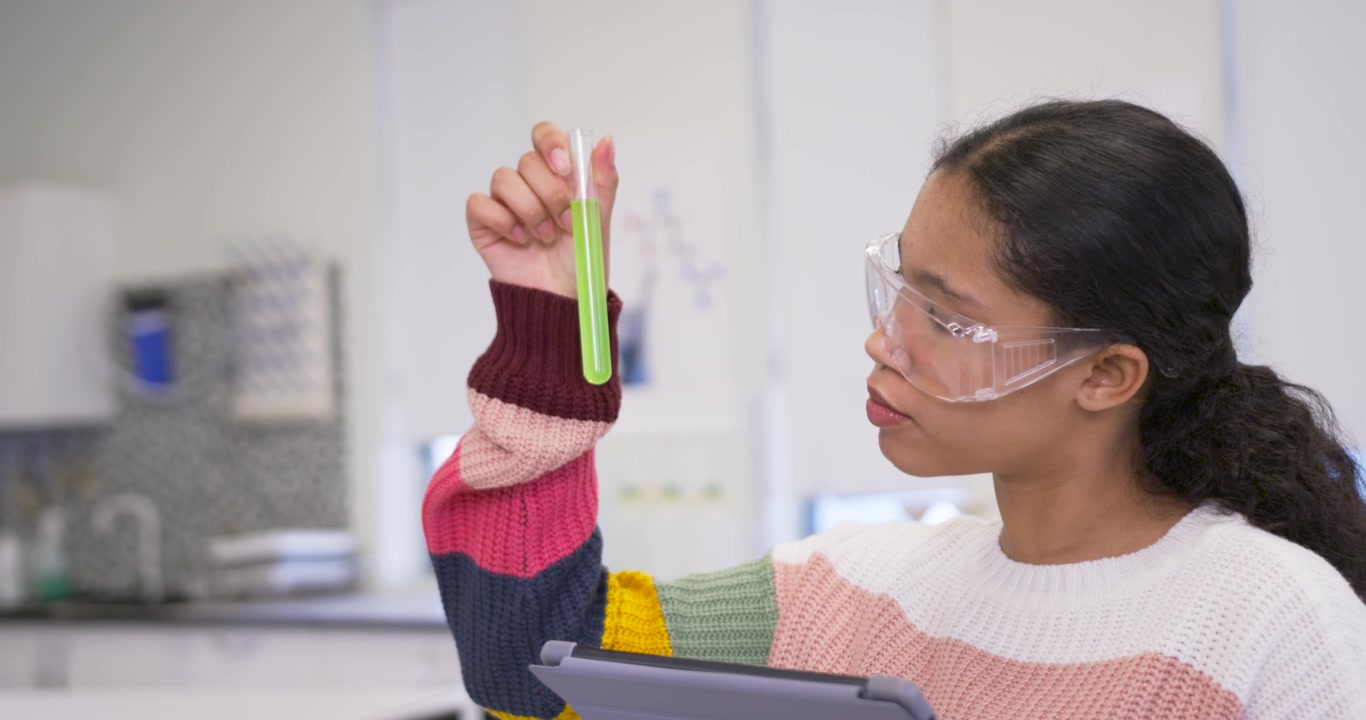 Schoolgirl in knit sweater with goggles holding test tube, checking tablet data in lab, copy space