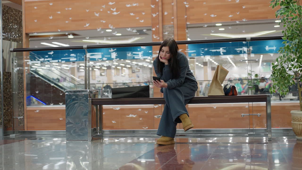 Young woman in dark outfit seated on bench inside shopping mall browses her phone with relaxed expression, shopping bags beside her on bench, surrounded by glass railing and potted plant