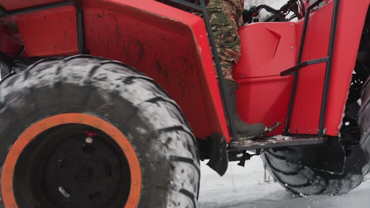 ATV rosso in un campo innevato, pilota con giacca mimetica e casco naviga un sentiero ghiacciato. Primo piano di pneumatici robusti che sollevano neve, vapore dallo scarico e dettagli del sottoscocca, portapacchi posteriore e sfondo boschivo.